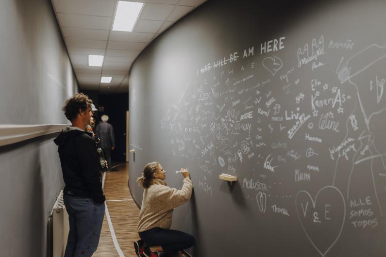 Ein Flur mit einer beschrifteten Wand: Eine Person kniet und schreibt mit Kreide oder Marker, während andere zuschauen. Die Wand ist mit vielen Namen und Zeichnungen bedeckt.