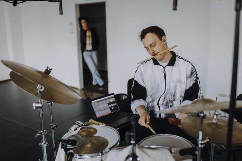 A drummer plays in a rehearsal room while holding a drumstick in his mouth. Another person stands in a doorway in the background.