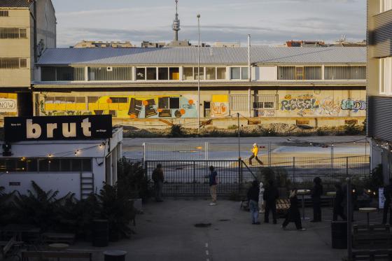 View from the brut courtyard towards an industrial building opposite with colourful graffiti. Several people stand in the foreground while a person in a yellow jumper walks across the site.