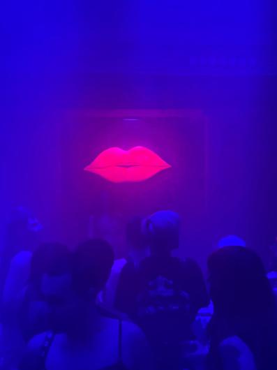 Ein dunkler, clubähnlicher Raum ist in intensives blau-violettes Licht und Nebel getaucht. In der Bildmitte leuchtet ein großes, neonpinkes Lippenmotiv, leicht geöffnet, wie ein schwebendes Symbol oder eine Lichtinstallation an der Wand oder Bühne. Die Lippen sind das hellste Element im Raum und ziehen den Blick sofort auf sich. Im Vordergrund sind mehrere Personen von hinten oder seitlich zu sehen. Ihre Gesichter sind nicht erkennbar, die Körper erscheinen als dunkle Silhouetten im farbigen Licht. Einige Personen tragen ärmellose Oberteile oder Tops; Frisuren und Details sind nur schemenhaft sichtbar. Die Menschen stehen dicht beieinander und blicken in Richtung der leuchtenden Lippen. Die Atmosphäre wirkt nächtlich, sinnlich und kollektiv, geprägt von Licht, Nebel und der Präsenz eines Publikums, wie bei einer Party, Performance oder Clubnacht.
