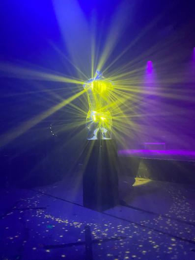 In a dark, fog-filled space, a torso-shaped sculpture without head or arms stands on a black pedestal. The surface appears reflective or faceted, resembling glass or metal. From inside the sculpture, an intense yellow light radiates outward, forming fan-shaped beams that spread through the room. The surrounding space is filled with blue and purple stage lighting, creating a strong contrast with the yellow glow. On the floor, small, scattered points of light appear, resembling stars or confetti. In the background, vague stage elements and haze are visible, with details remaining indistinct. The overall impression is atmospheric, futuristic, and performative, evoking a light installation or art object within a club night or performance setting.
