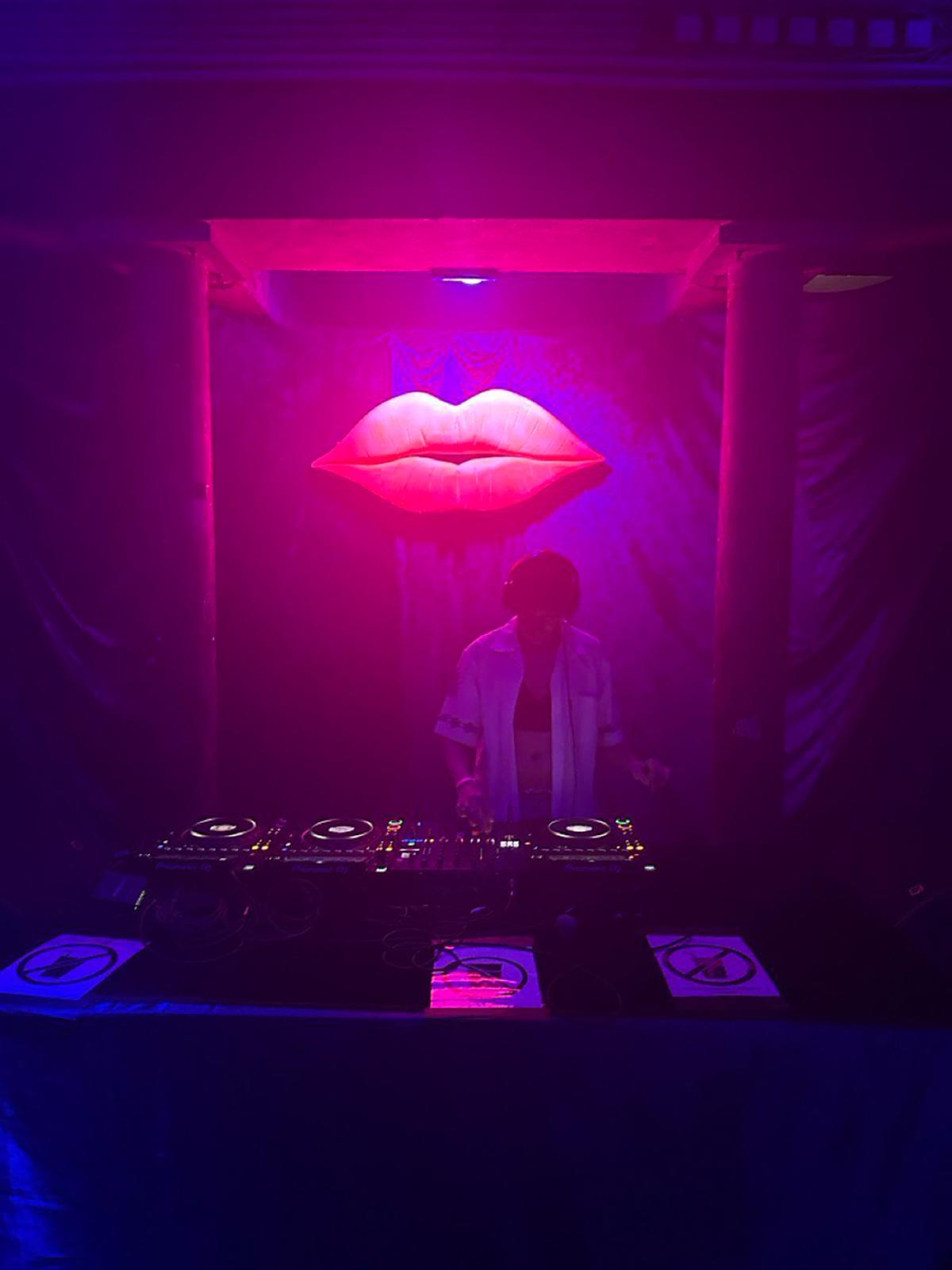 In einem dunklen, nebeligen Raum steht eine DJ-Person hinter einem DJ-Pult mit zwei Plattenspielern und einem Mischpult. Die Person trägt Kopfhörer und ein offenes, helles Hemd über einem Oberteil; das Gesicht ist durch Gegenlicht und Nebel nur schemenhaft sichtbar. Über dem DJ-Pult hängt mittig eine große, leuchtend pinke Lippen-Skulptur, die an einer Wand befestigt ist. Die Szene ist in violettes und magentafarbenes Licht getaucht, das den Raum weich ausleuchtet und die Konturen verschwimmen lässt. Das DJ-Pult steht zwischen zwei massiven Säulen, die den Raum rahmen. Die Atmosphäre wirkt clubartig, sinnlich und performativ, mit Fokus auf Licht, Sound und Präsenz.