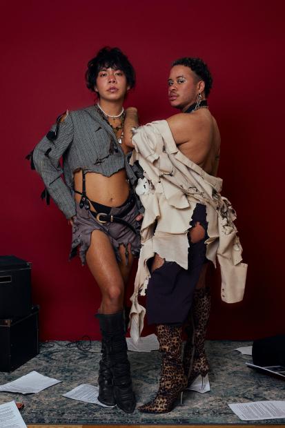 Zwei Personen stehen dicht nebeneinander vor einem tiefroten Hintergrund und blicken direkt in die Kamera. Beide tragen bewusst zerrissene, dekonstruktive Kleidung in gedeckten Farben. Die Person links trägt ein kurzes, aufgerissenes Oberteil und eine kurze Hose mit freiliegendem Bauch, dazu hohe dunkle Stiefel. Ketten und Schmuck liegen um Hals und Oberkörper. Die Haltung wirkt selbstbewusst, mit leicht angewinkeltem Bein und offener Körperausrichtung. Die Person rechts trägt ein asymmetrisches, helles Kleidungsstück mit offenen Schultern und freiliegendem Rücken, kombiniert mit hohen Stiefeln mit Leopardenmuster. Der Körper ist leicht zur Seite gedreht, ein Arm liegt auf der Schulter der anderen Person. Auf dem Boden liegen verstreute Papierblätter und Kabel, im Hintergrund sind dunkle technische Objekte sichtbar. Das Bild wirkt inszeniert, kraftvoll und intim, mit Fokus auf Körperhaltung, Materialität der Kleidung und die Beziehung zwischen den beiden Personen.
