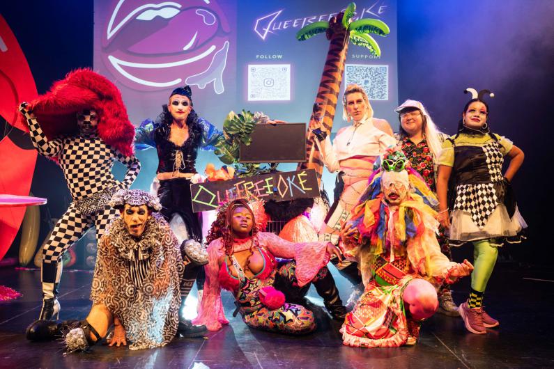 Gruppenporträt auf einer Bühne: Mehrere Personen in bunten, extravaganten Kostümen und Make-up posieren um einen dekorierten Karaoke-Stand. Im Hintergrund sind eine große Lippen-Grafik, der Schriftzug Queereeoké und zwei QR-Codes zu sehen. Bunte Bühnenlichter und Requisiten 