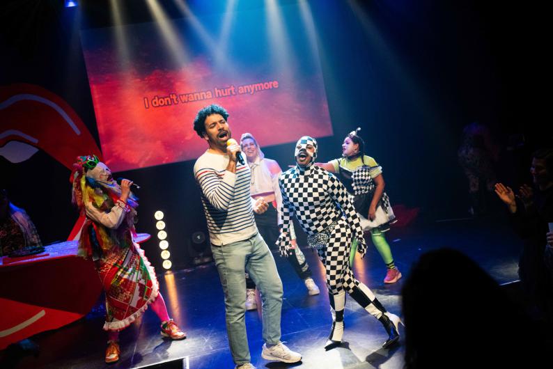 Bühnenszene bei einer Karaoke-Performance: Eine Person in gestreiftem Oberteil singt mit Mikrofon im Vordergrund, umgeben von mehreren kostümierten Performer*innen. Auf der Leinwand dahinter steht der Liedtext „I don’t wanna hurt anymore“. Blaues und rotes Licht, Bewegung und Publikum am Rand vermitteln Konzert- und Party-Stimmung.