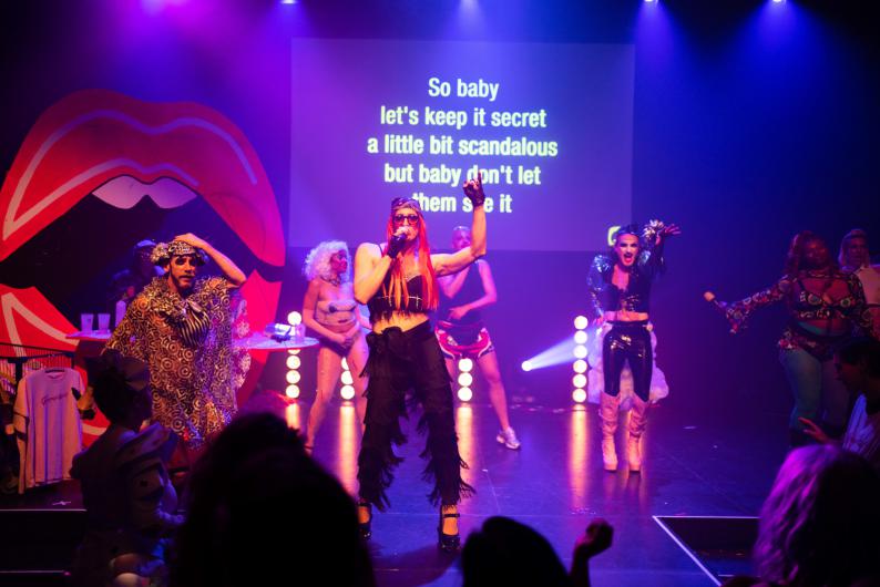 Mehrere Personen performen auf einer Bühne unter violettem Licht. Im Zentrum singt eine Person mit langem rotem Haar und Mikrofon, während andere Personen in auffälligen Outfits tanzen oder mitsingen. Auf der Leinwand im Hintergrund läuft Karaoke-Text in großen gelben Buchstaben.