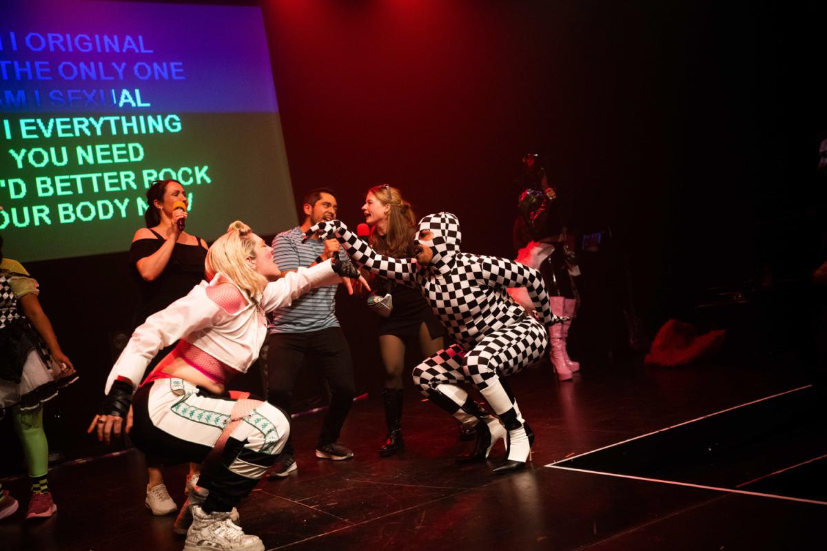 Karaoke-Moment auf einer rot beleuchteten Bühne: Zwei Personen aus dem Publikum singen mit Mikrofonen, während eine Person im schwarz-weißen Schachbrettkostüm sich dynamisch zu ihnen beugt. Weitere Performer*innen und Karaoke-Text auf einer Leinwand sind im Hintergrund sichtbar. Die Szene wirkt spontan, laut und spielerisch.