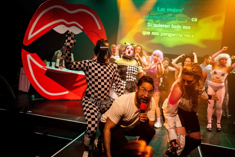 Eine Gruppe von Performer*innen und Teilnehmenden steht dicht vor der Bühne und singt gemeinsam in Mikrofone. Auffällige Kostüme, warmes gelb-oranges Licht und eine grosse rote Lippen-Requisite prägen das Bild. Auf der Leinwand im Hintergrund ist spanischer Karaoke-Text zu lesen.
