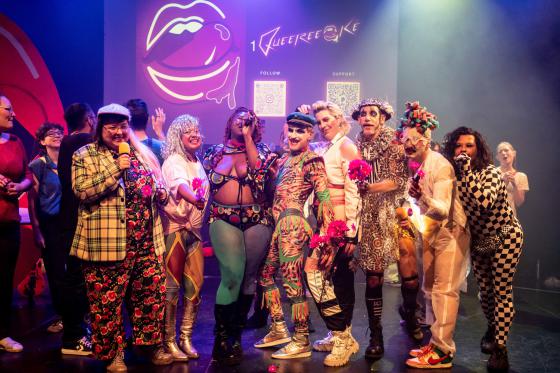 Gruppenbild auf einer Bühne nach oder während einer Performance: Mehrere Personen in auffälligen, queer-futuristischen oder glamourösen Kostümen stehen eng zusammen und halten pinke Blumen in die Kamera. Im Hintergrund sind das Queereeoké-Logo, eine Lippen-Grafik und zwei QR-Codes projiziert. Das Bild wirkt feierlich, kollektiv und verspielt.