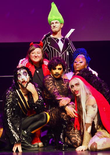 Gruppenporträt von sechs Personen auf einer Bühne, inszeniert wie ein Show-Finale. Die Personen tragen auffällige Kostüme, Make-up und Perücken in kräftigen Farben, darunter Glitzer, Spitze und ein leuchtend grüner, hochgestylter Haarschopf. Einige Personen knien oder sitzen im Vordergrund, andere stehen dahinter. Die Posen sind selbstbewusst, spielerisch und expressiv. Violettes Bühnenlicht verstärkt die theatralische, queere Ästhetik der Szene.
