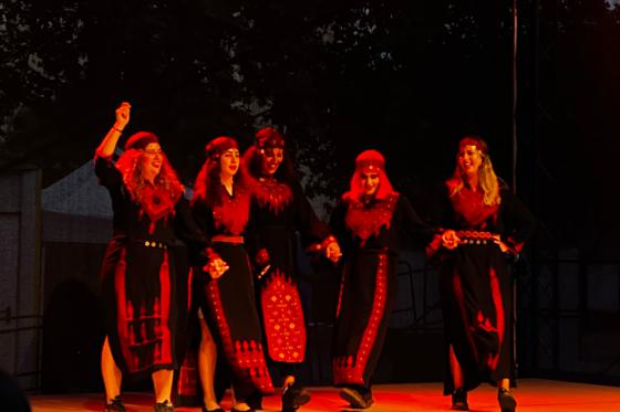 Five people stand on an outdoor stage holding hands. They wear long black garments with red patterns and matching head coverings. In warm red stage light they perform a dance movement together, with one person raising an arm. Dark trees are visible in the background.