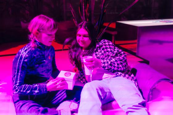 Zwei Personen sitzen entspannt nebeneinander auf einer Sitzlandschaft in pink-violettem Licht. Eine Person hält eine kleine Schachtel in den Händen und öffnet sie, während die andere Person einen Becher hält und aufmerksam hineinschaut. Beide wirken in ein ruhiges, gemeinsames Gespräch vertieft. Im Hintergrund sind Pflanzen und Möbel angedeutet, die Szene erinnert an eine Lounge- oder Pausensituation in einem künstlerischen Umfeld.
