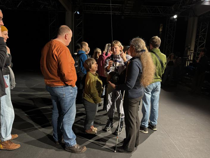 Auf einer dunklen Theaterbühne steht eine Gruppe von Besuchenden in Alltagskleidung zusammen. Im Zentrum erklärt eine Person in grauem Overall etwas und hält ein kleines Gerät mit Kabeln; davor hält eine andere Person eine große schwarze Kopfhörer-/Audiogeräte-Einheit und stützt sich auf einen weißen Langstock. Scheinwerfer und Bühnentraversen sind im Hintergrund zu sehen.