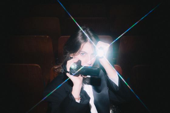 A person sits in a dark cinema between brown wooden seats, pointing a small video camera with a bright light towards the viewer, light beams crossing the image in a star shape.