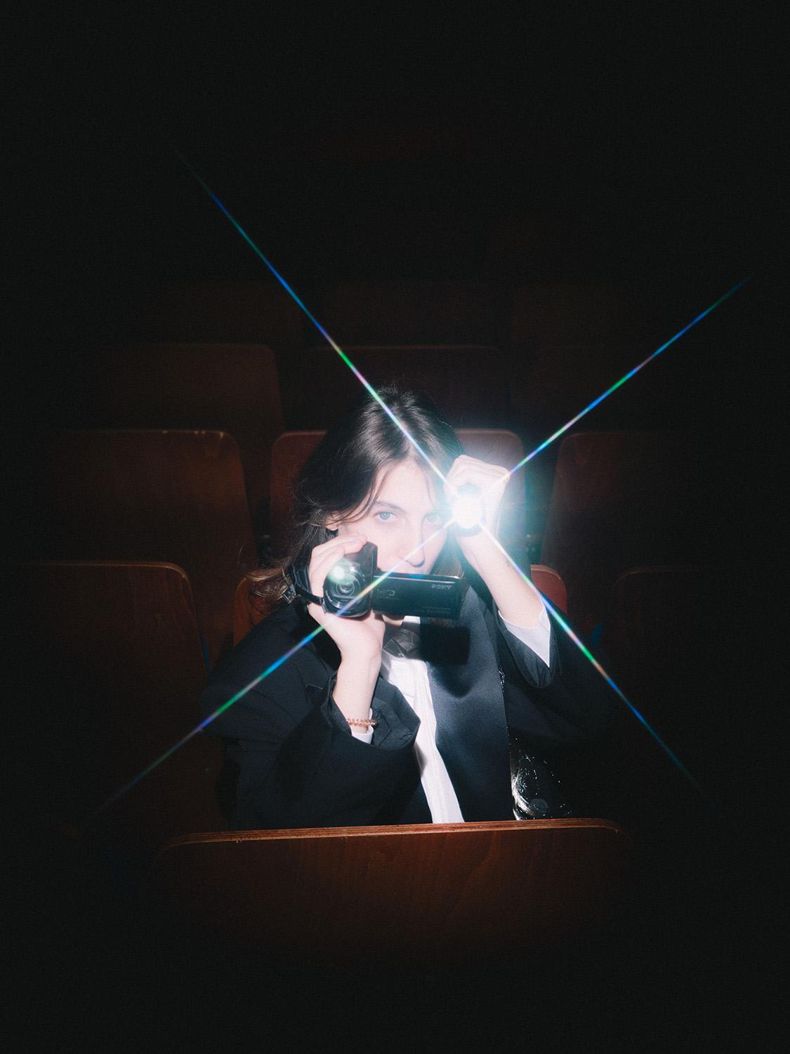 A person sits in a dark cinema between brown wooden seats, pointing a small video camera with a bright light towards the viewer, light beams crossing the image in a star shape.