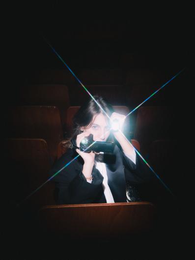 A person sits in a dark cinema between brown wooden seats, pointing a small video camera with a bright light towards the viewer, light beams crossing the image in a star shape.