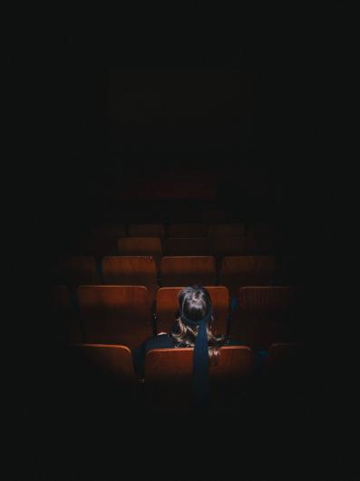 Rear view of a person with long hair and a wide dark ribbon, sitting alone in a lit seat in an otherwise dark cinema and facing the empty screen.