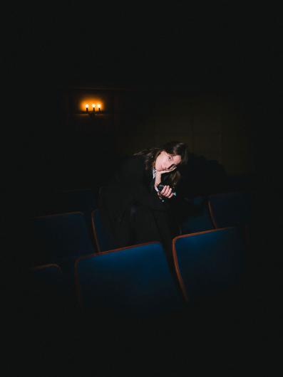 A person dressed in black leans across several blue cinema seats, looking directly at the camera while holding a small recording device, with two warm wall lights glowing in the background.