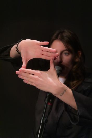 A performer stands at a microphone, forming a rectangular frame with both hands in front of their face. The background is dark, and the face is partially obscured.