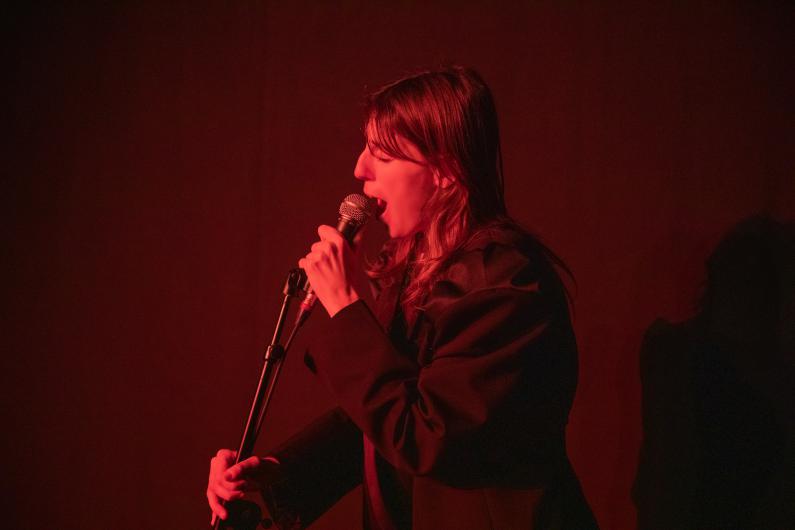 Eine Person singt in ein Mikrofon, beleuchtet von rotem Bühnenlicht. Der Hintergrund ist dunkel, die Szene wirkt konzentriert und intim.