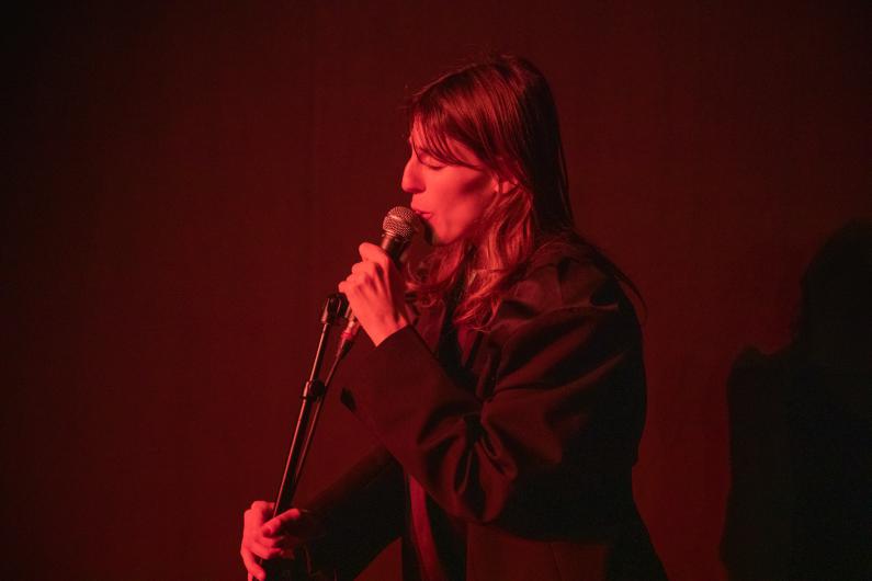 A performer sings into a microphone under red stage lighting. The background is dark, creating a focused and intimate atmosphere.
