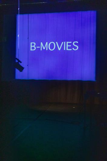 A stage with a large projection screen displaying the text “B-MOVIES” in bright letters on a blue background. The space is dark, with technical equipment visible at the edges.