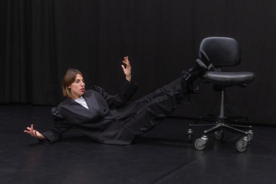 Eine Person liegt auf dem Bühnenboden und lehnt sich seitlich auf den Arm. Die Beine ruhen auf einem schwarzen Bürostuhl mit Rollen. Die Person trägt einen dunklen Anzug.
