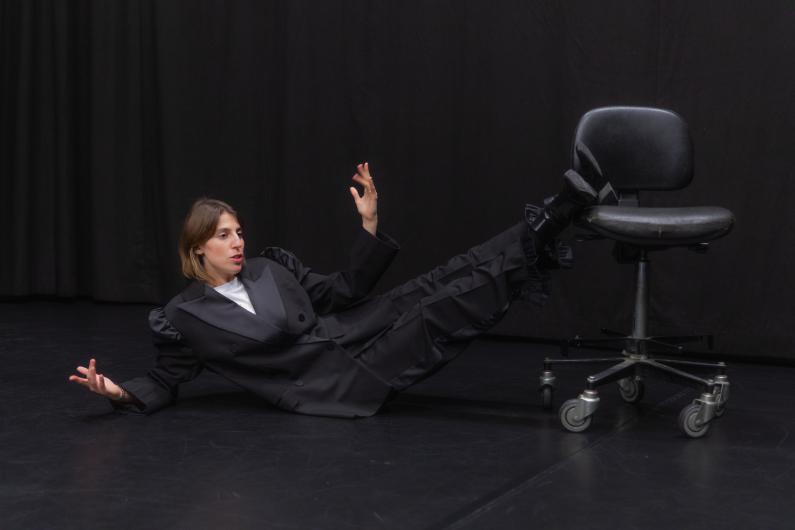 Eine Person liegt auf dem Bühnenboden und lehnt sich seitlich auf den Arm. Die Beine ruhen auf einem schwarzen Bürostuhl mit Rollen. Die Person trägt einen dunklen Anzug.