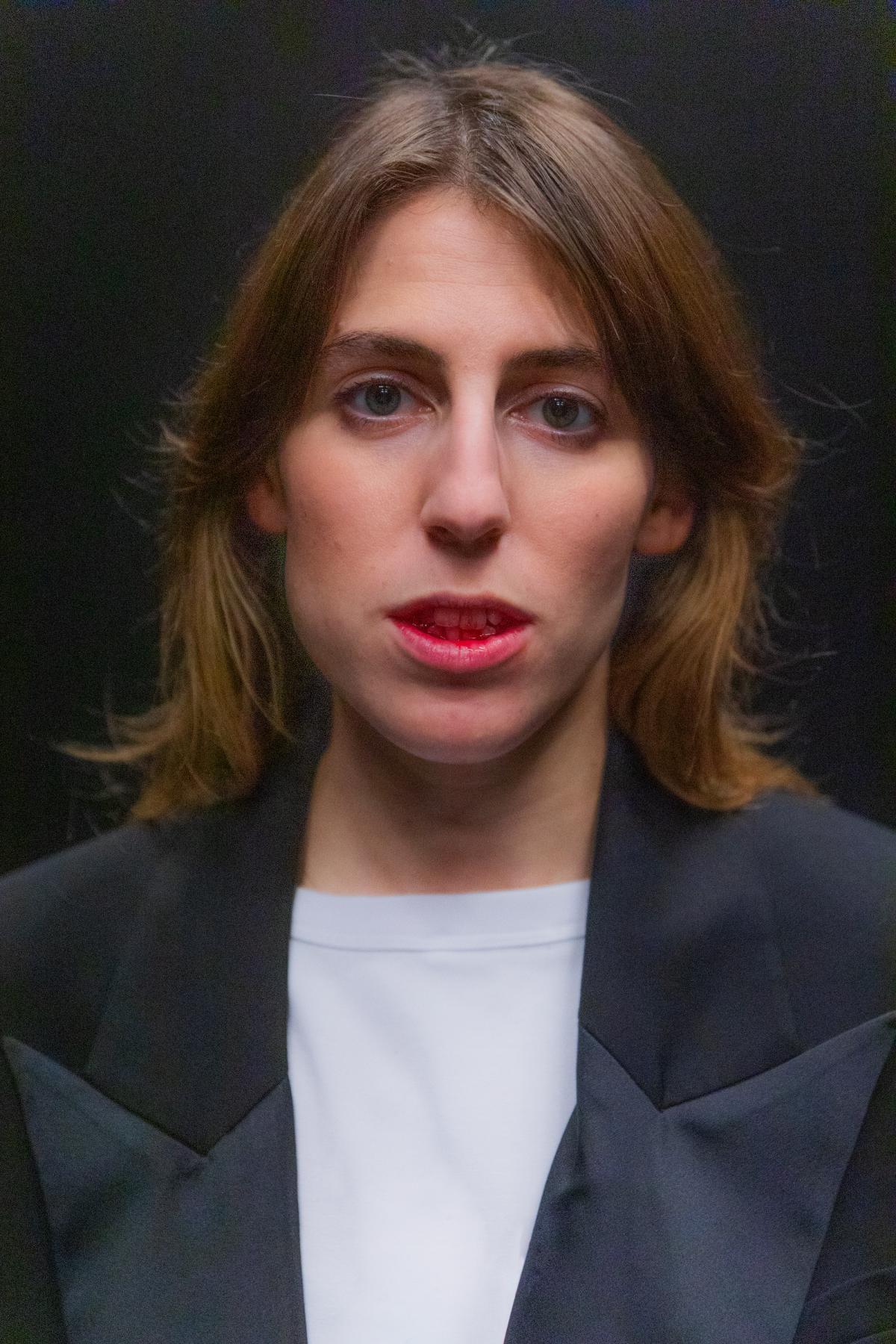 Portrait of a person with short light brown hair against a dark background, facing forward. The person wears a dark jacket over a light top.