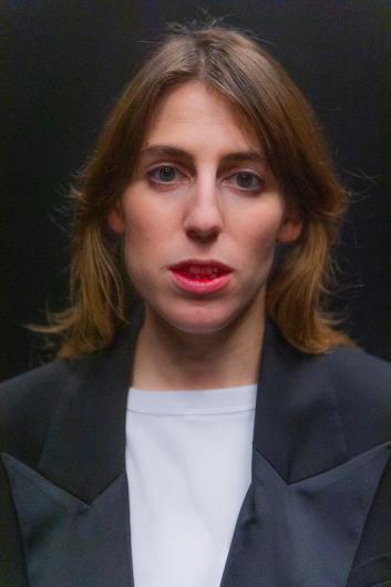 Portrait of a person with short light brown hair against a dark background, facing forward. The person wears a dark jacket over a light top.
