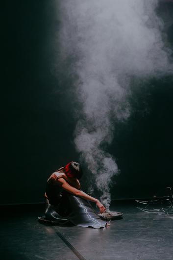 A performer sits on the floor of a dark stage beside a shallow metal bowl emitting smoke. Their body is folded inward as the smoke rises upward.