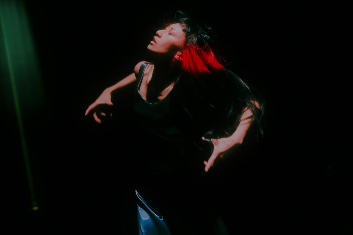 A performer with long dark hair with red streaks moves on a dark stage under a spotlight, head tilted back with arms open in an expressive movement.