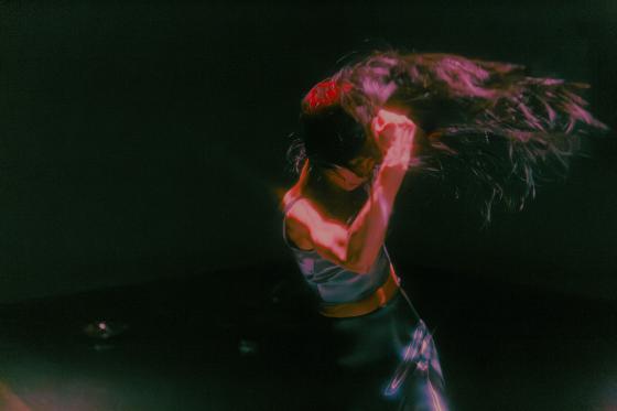 A performer swings their head forward, tossing long hair in a fast movement. Stage lighting creates glowing color halos around the body and hair.