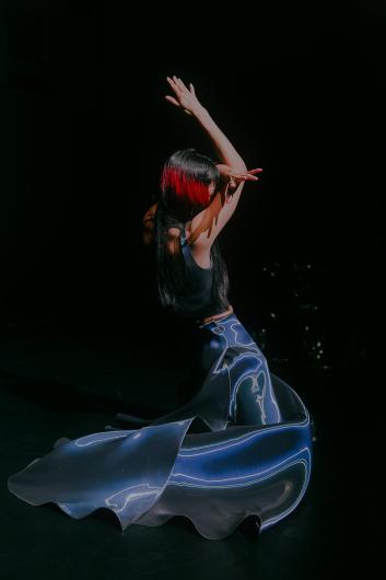 Eine Performer*in mit langen schwarzen Haaren und roten Strähnen kniet auf einer dunklen Bühne im Spotlicht. Die Arme sind nach oben erhoben und bilden eine expressive, tänzerische Geste. Ein langes, glänzendes Kostüm breitet sich in weichen Wellen um den Körper auf dem Boden aus und reflektiert das Licht.