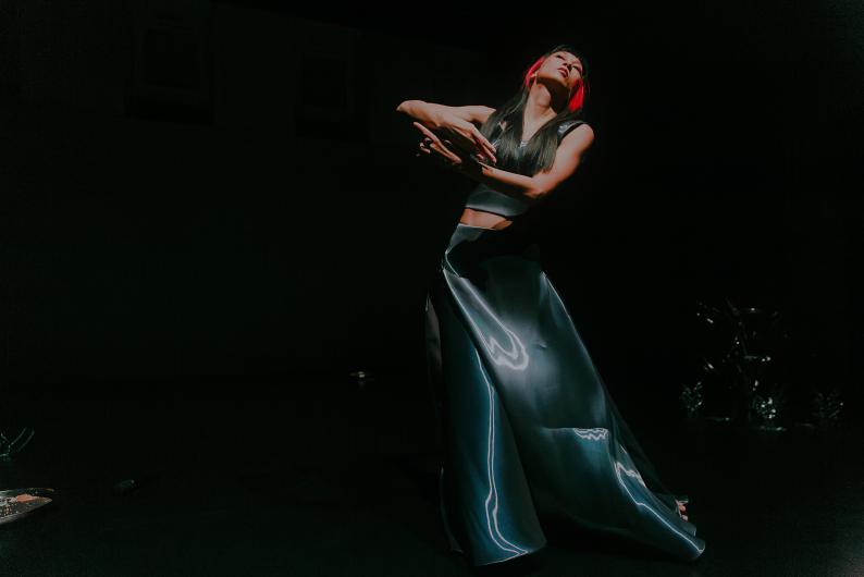 A performer with long black hair and red streaks moves on a dark stage under a spotlight. Their body leans slightly back while the arms cross in a flowing gesture. A long reflective costume catches the light and emphasizes the turning movement.