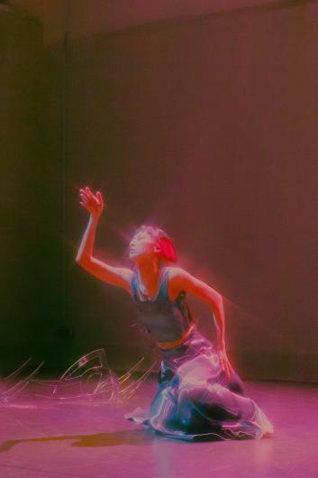 A performer kneels on the stage with one arm raised toward the light. Colored stage lighting creates soft rainbow reflections in the space.