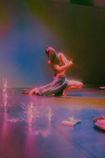 Eine Performer*in kniet auf dem Boden und dreht den Oberkörper in einer expressiven Pose. Auf dem Boden liegen glänzende transparente Objekte, die das Licht reflektieren.