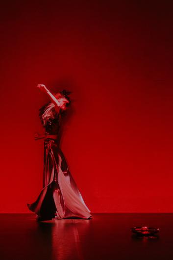 A performer moves across a stage in front of a bright red background. Their body leans slightly back with one arm raised while their shiny costume reflects the light.