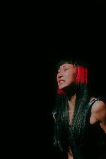 Nahaufnahme einer Performer*in mit langen schwarzen Haaren und roten Strähnen auf einer dunklen Bühne. Das Gesicht ist vom Spotlicht beleuchtet, die Augen sind geschlossen und der Ausdruck wirkt intensiv und emotional.
