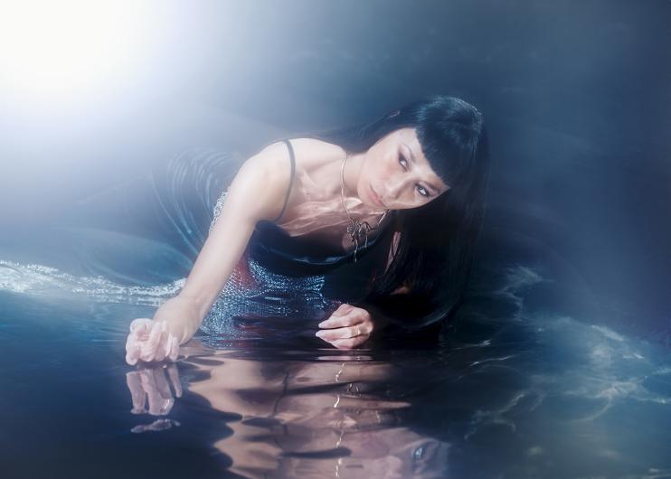 Die Performer*in Cat Jimenez liegt seitlich in einem dunklen, nebligen Raum am Boden und stützt sich mit einem Arm auf, während die andere Hand die spiegelnde Wasseroberfläche berührt; das lange dunkle Haar fällt nach vorne, ein Lichtschein links oben überstrahlt den Bildrand und lässt Körper und Wasser weich schimmern.