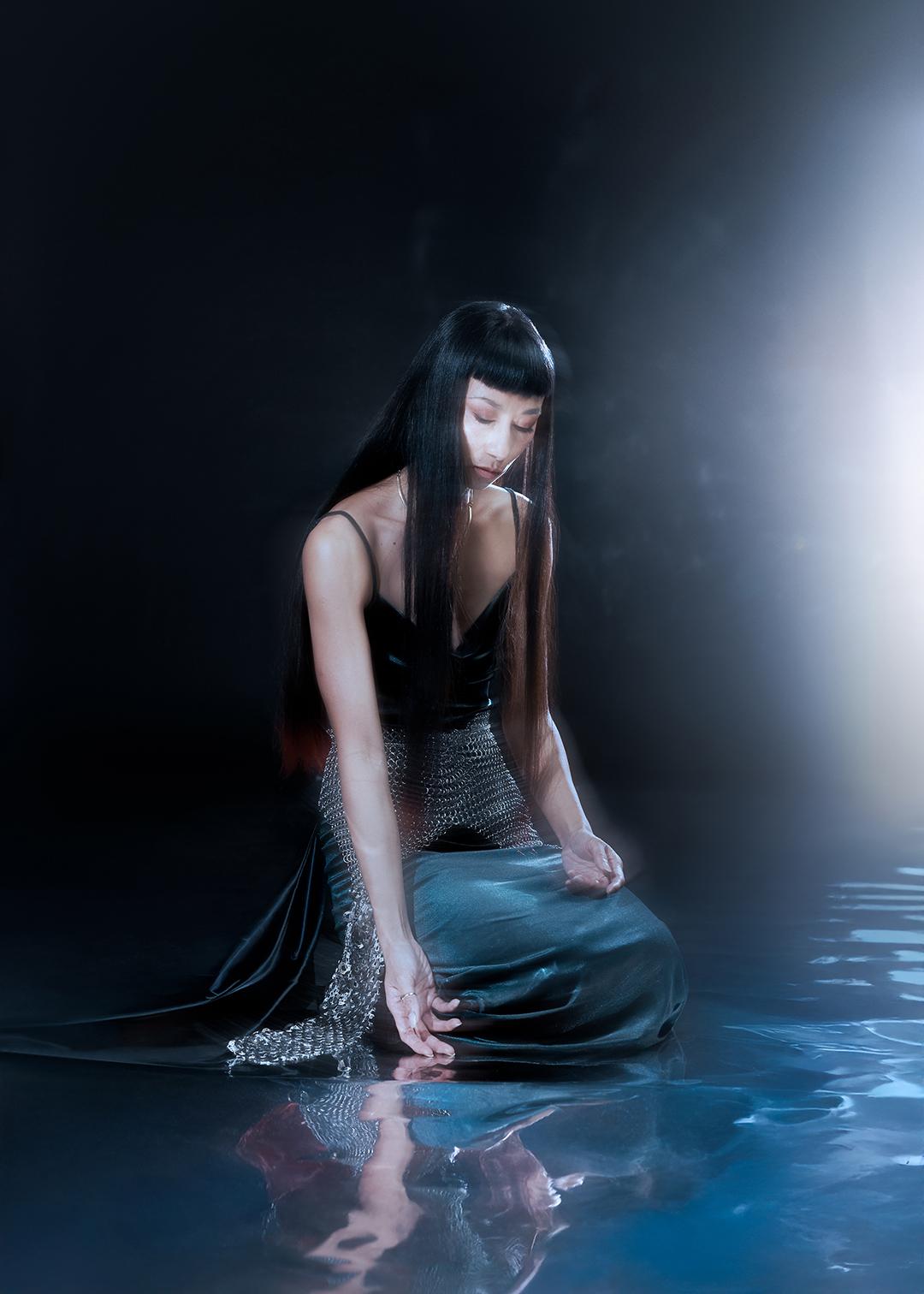 Cat Jimenez kneels in a dark space at the edge of a pool of water, looking down with one hand resting in the water; their long straight hair falls over the shoulders, a fitted dark dress and a shimmering chain or sequin element reflect on the surface while a bright beam of light enters the image from the right.