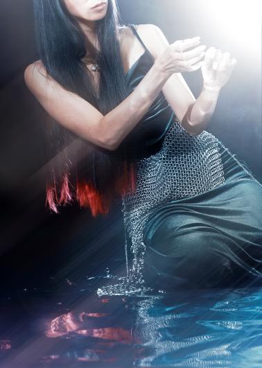 Nahaufnahme von Cat Jimenez’ Oberkörper in einem dunklen Kleid mit feinem Kettenelement; der Kopf ist nur teilweise zu sehen, die Arme sind nach vorne gehoben und fangen einen intensiven Lichtschein rechts im Bild ein, während sich die Figur im bewegten Wasser am Boden spiegelt.