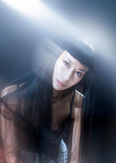 Portrait of Cat Jimenez with long straight hair and blunt fringe in a dark top, body leaning slightly forward; a diagonal beam of light from the upper left cuts across the image, softly illuminating face and shoulders against a dark background.