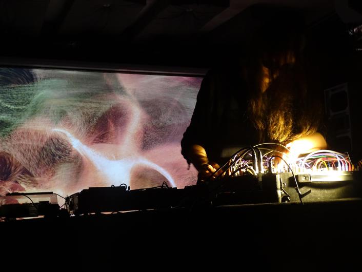 Eine Person steht in einem dunklen Raum hinter einem Tisch mit elektronischem Musik-Equipment. Vor der Person befindet sich ein Modular-Synthesizer mit vielen Kabeln, Knöpfen und kleinen Lichtern, die hell leuchten. Die Hände der Person liegen am Gerät und scheinen Einstellungen zu verändern. Das Gesicht ist nicht sichtbar; lange Haare fallen nach vorne und werden von einem warmen Licht angestrahlt. Im Hintergrund ist eine großflächige Projektion zu sehen: abstrakte, organische Formen in weichen Rosa-, Beige- und Grüntönen, die an Haut, Wellen oder Bewegung erinnern. Die Szene wirkt konzentriert, intim und atmosphärisch, wie bei einer Live-Performance.