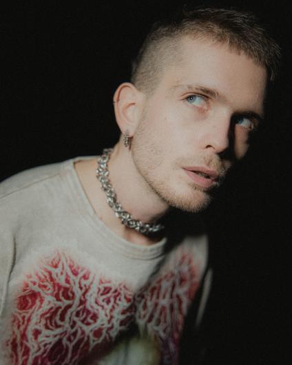 Close-up of a person against a dark background. The face is lit from the side, with short hair, visible earrings, and a chain necklace. They wear a light-colored T-shirt with a red, organic-looking print and look slightly to the side.