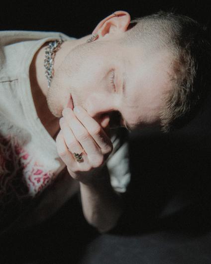 Close-up of a person against a dark background. The person tilts or rests their head to the side with eyes closed. One hand with a ring is held close to the face; short hair, an earring, and a chain necklace are visible. Soft side lighting creates a quiet, intimate mood.
