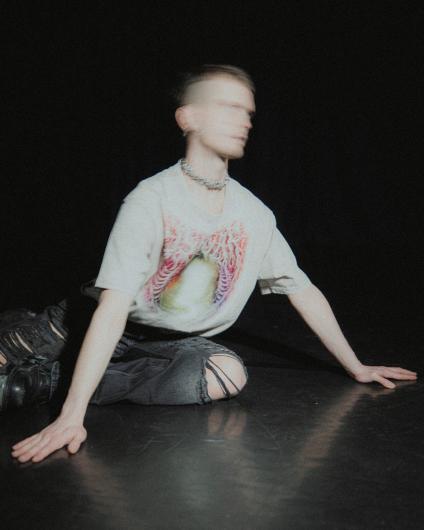 A person sits on the floor against a dark background, legs folded to the side and hands resting on the ground. The head is in motion and slightly blurred. The person wears a light T-shirt with a graphic print, a chain necklace, and dark ripped jeans. Low light creates a focused, physical mood.