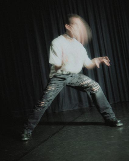 Eine Person steht in breiter Haltung vor dunklem Vorhang. Der Kopf ist stark verwischt durch Bewegung, die Arme sind angewinkelt und aktiv. Helles T-Shirt, Kette und zerrissene dunkle Jeans. Gedämpftes Licht, dynamische Szene.