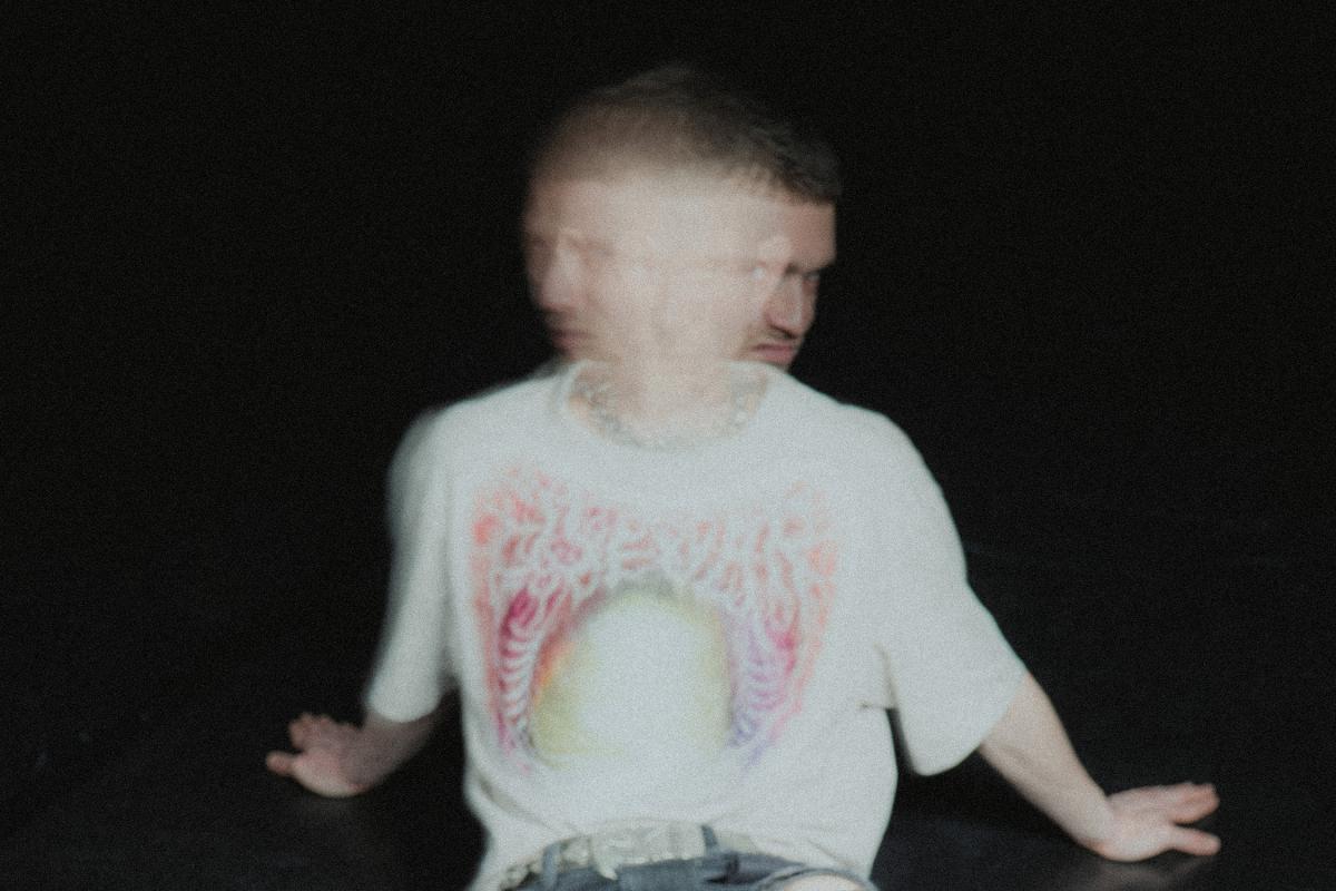 A person sits against a black background, leaning back on both hands. Heavy motion blur makes the face appear doubled, like two overlapping profiles. They wear a light T-shirt with a colorful print and a chain necklace.