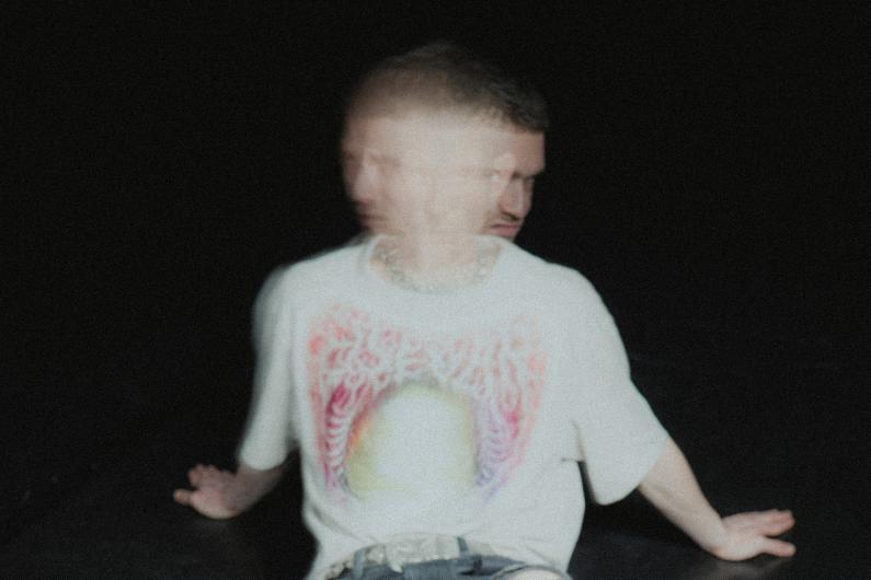 A person sits against a black background, leaning back on both hands. Heavy motion blur makes the face appear doubled, like two overlapping profiles. They wear a light T-shirt with a colorful print and a chain necklace.